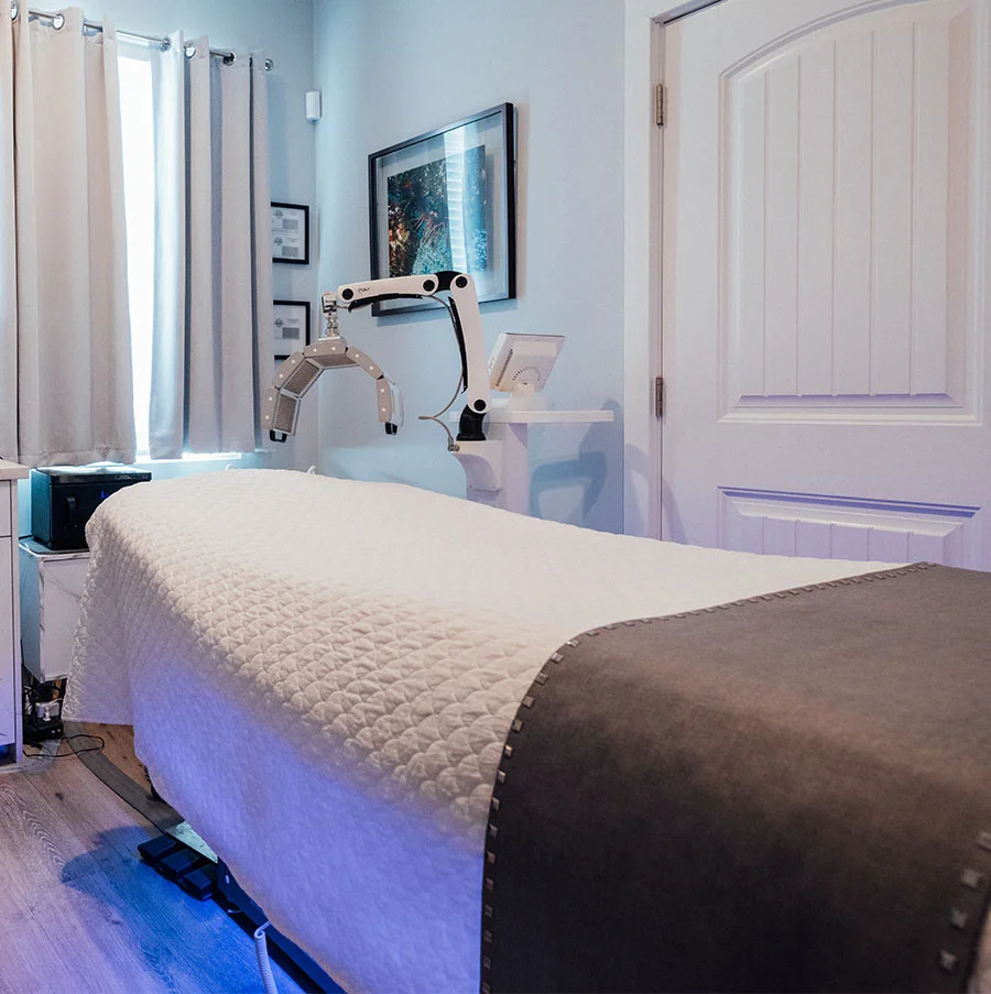 The image shows a treatment room corner with a white massage table, medical equipment, pink curtains, and framed pictures on blue walls. A door is on the right, and purple-blue lighting washes over the light wood floor. - Aqua Gold in San Jose, CA