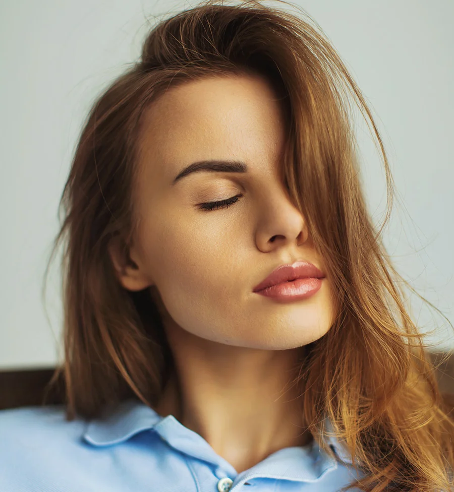 Close-up of a woman with shoulder-length, light brown hair in loose waves. A strand falls across her forehead as her eyes remain closed, creating a serene expression. Her smooth, light beige skin and soft pinkish-nude lips are gently highlighted by soft, natural indoor light. She wears a light powder-blue collared shirt against a muted gray-beige background. - Aqua Gold Canidates in San Jose, CA