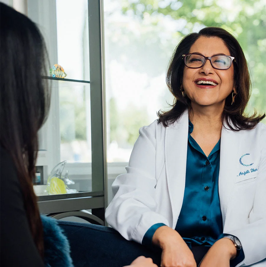 The image shows a middle-aged woman with shoulder-length brown hair, wearing a white lab coat over a teal silk shirt. She has gold hoop earrings, a watch on her left wrist, and is smiling warmly at someone seated across from her, partially visible in the frame. Behind her are glass shelves and a large window with trees outside, suggesting a bright, modern office setting on a sunny day. - Bio Identical Hormone Therapy in San Jose, CA