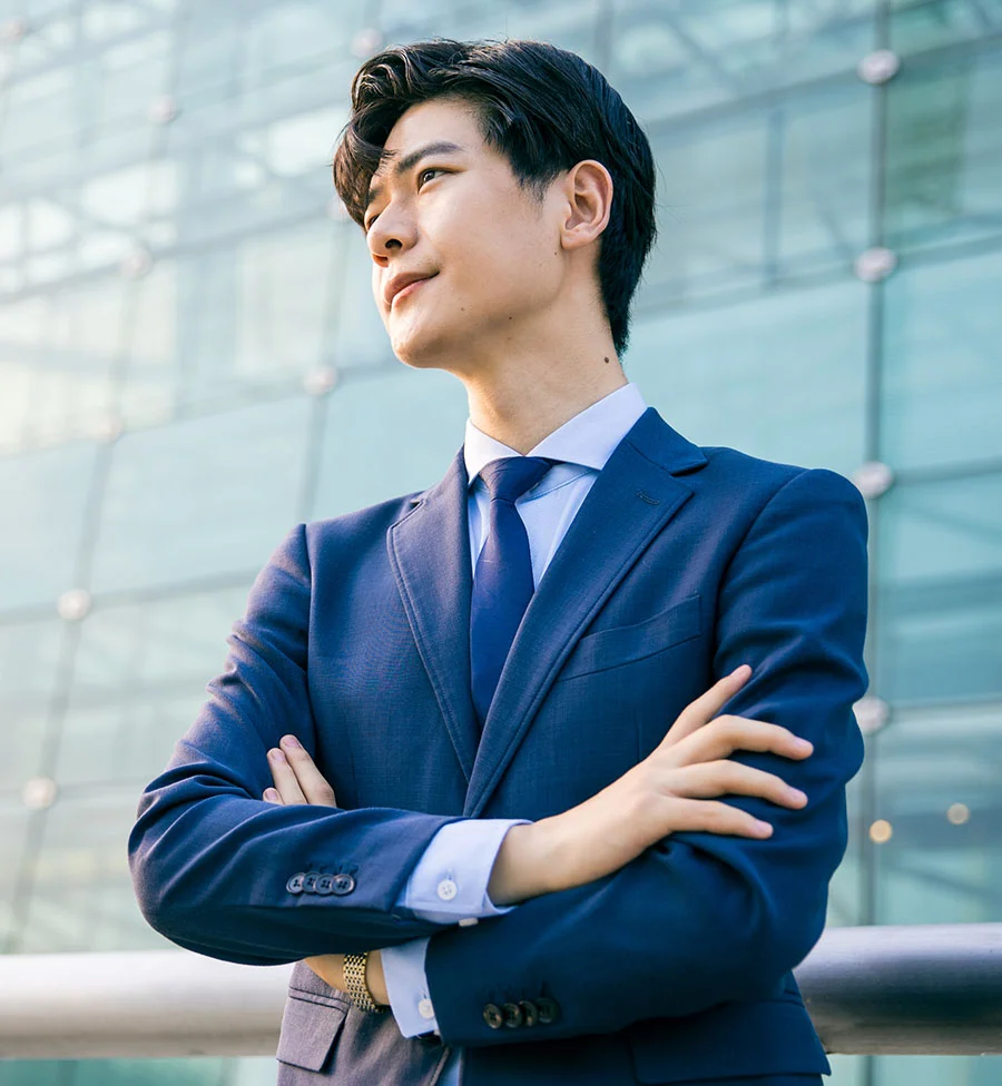The image shows a well-dressed Asian man standing confidently in front of a modern glass building. He wears a blue suit with a white shirt and blue tie, and his dark, neatly styled hair adds to his professional appearance. With arms crossed, he stands before large glass panels and metal frameworks, the blurred background hinting at a busy urban setting. - Bio Identical Hormone Therapy for Men in San Jose, CA