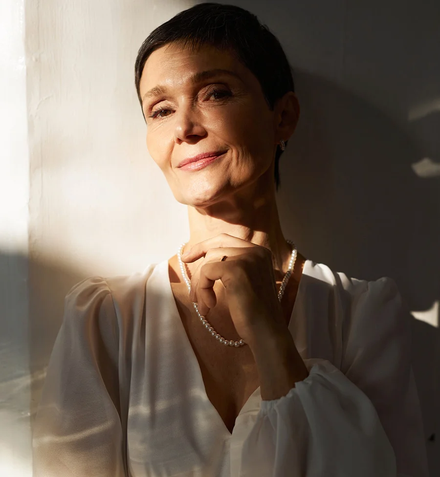 The image shows a mature woman with short dark brown hair, angled slightly toward the camera and gazing directly at the viewer with a serene, subtle smile. She wears an off-white V-neck blouse and a delicate pearl necklace, her right hand resting gently under her chin. Sunlight casts soft shadows across the plain white background, highlighting her natural glow and the texture of her clothing, creating an elegant and tranquil mood. - Bio Identical Hormone Therapy in San Jose, CA