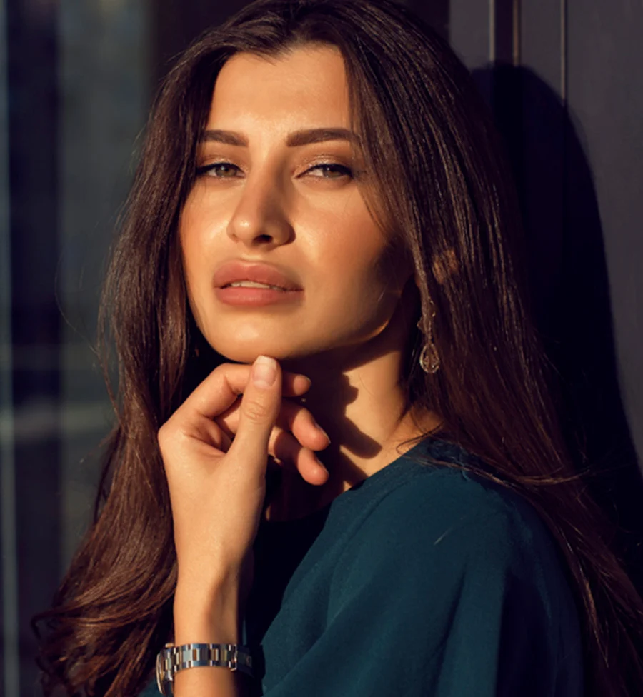 A portrait of a woman with long brown hair wearing a teal shirt, a silver-tone watch, and dangling silver earrings. She holds her wrist gracefully, drawing attention to her hand. Behind her is a blurred urban backdrop with a large window. Angled light from the right highlights her skin, lips, and hair, creating a striking contrast against the surrounding shadows. The composition’s triangular light and dark areas give the image a stylish, contemplative feel. - Chemical Peel Canidates in San Jose, CA