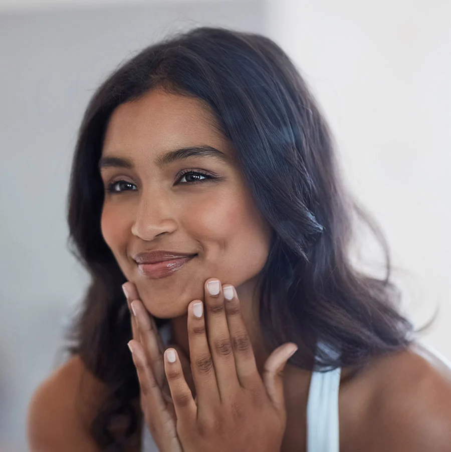 A smiling woman with long brown hair, wearing a light-colored top and rests her hands on her face as she smiles gently. A window is seen blurred in the background as she is looking directly at the camera. - Dermal Fillers Benefits in San Jose, CA
