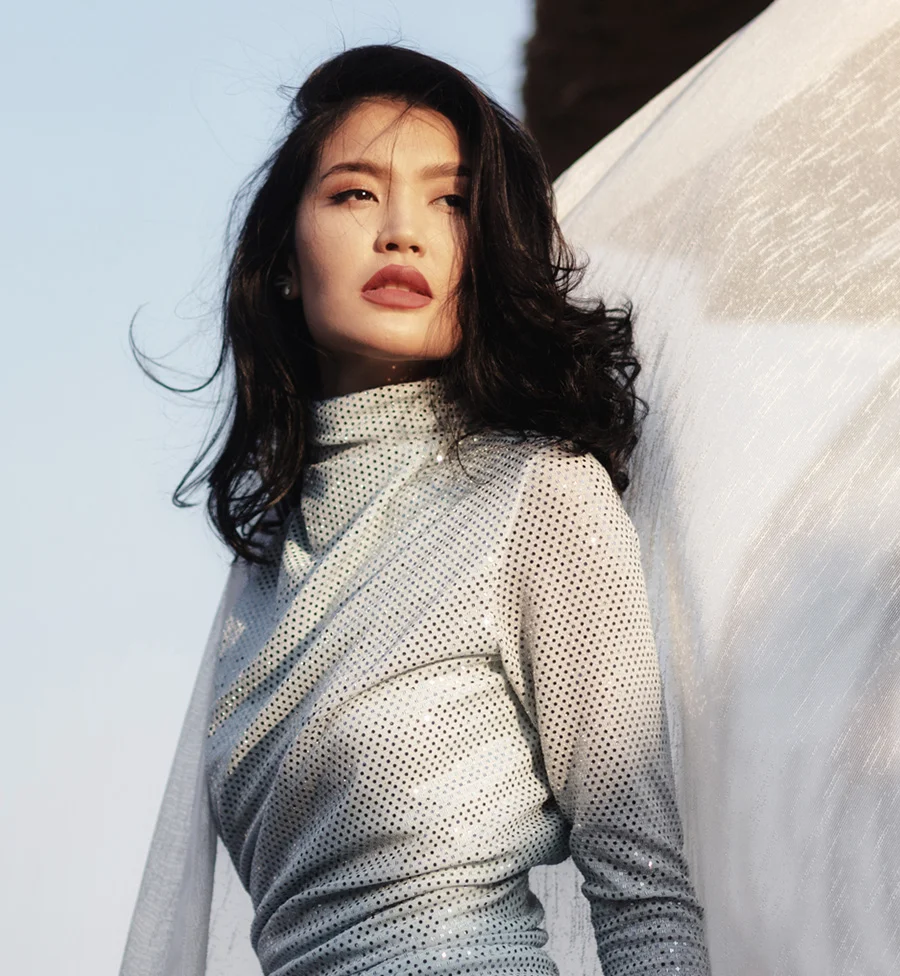 The image shows a striking portrait of a woman with dark, loose hair wearing a white, long-sleeved lycra dress detailed with ruching, black rhinestones, and sheer chiffon accents. A zipper runs from the torso to the neck. She tilts her head slightly right, gazing directly at the viewer while leaning on translucent white fabric lit with a warm orange glow. The dark floor and blue sky background add contrast, creating an intriguing and dramatic atmosphere. - Dermal Fillers in San Jose, CA