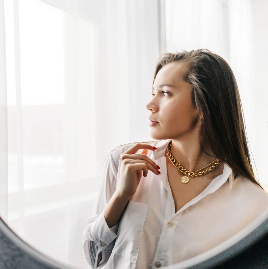 A young woman with long brown hair, wearing a white button-down shirt and a gold pendant necklace, is seen through a round mirror. Her hand rests at her chin as she gazes off to the right. White curtains form a clean, minimalist backdrop, creating a serene and contemplative mood. - Dermaplaning in San Jose, CA