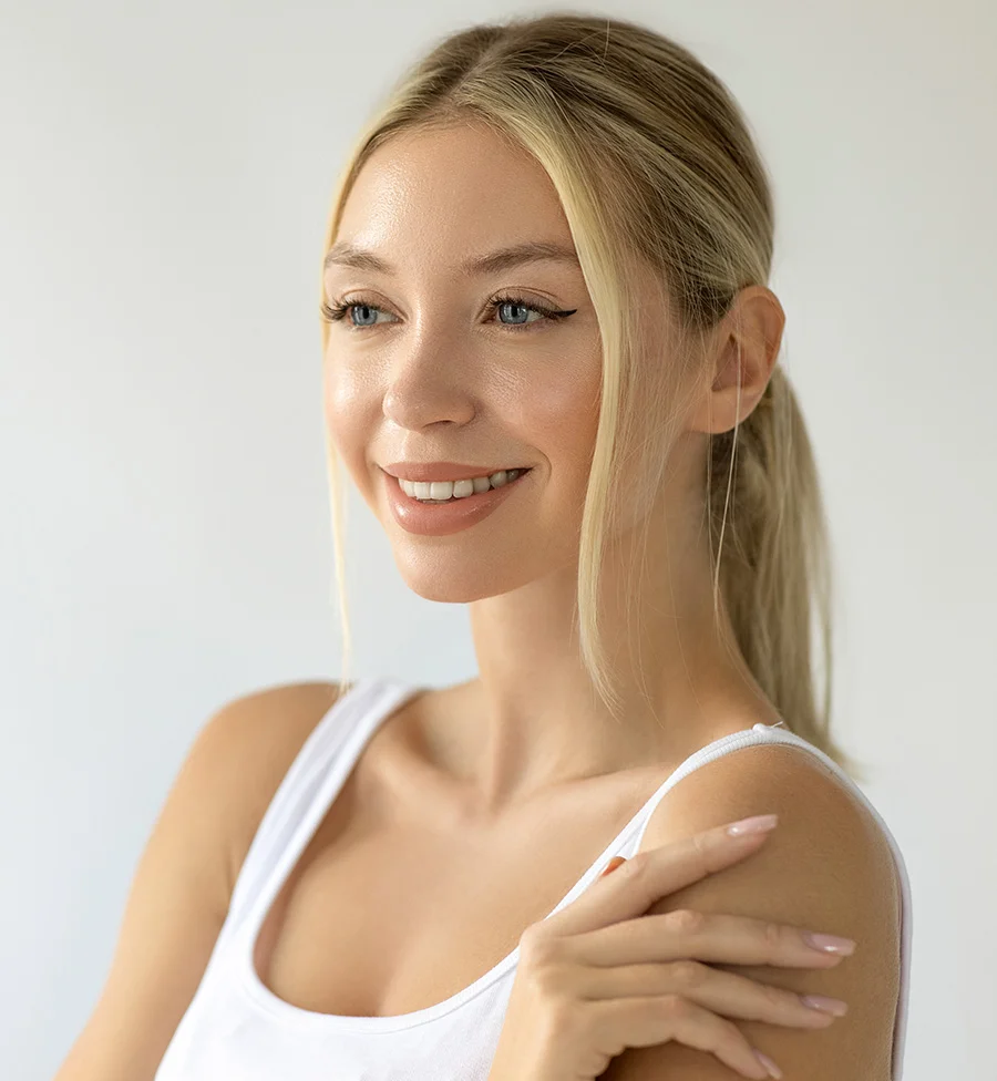 The image shows a blonde woman with her hair in a low ponytail, wearing subtle makeup and a white tank top. A few loose strands frame her face, and her blue eyes are highlighted by soft eyeliner. Her fair skin and neutral-polished nails complement her minimal look. She poses confidently with her right elbow resting on her left shoulder against a clean white background, exuding relaxed poise and natural beauty. - Dermaplaning Canidates in San Jose, CA