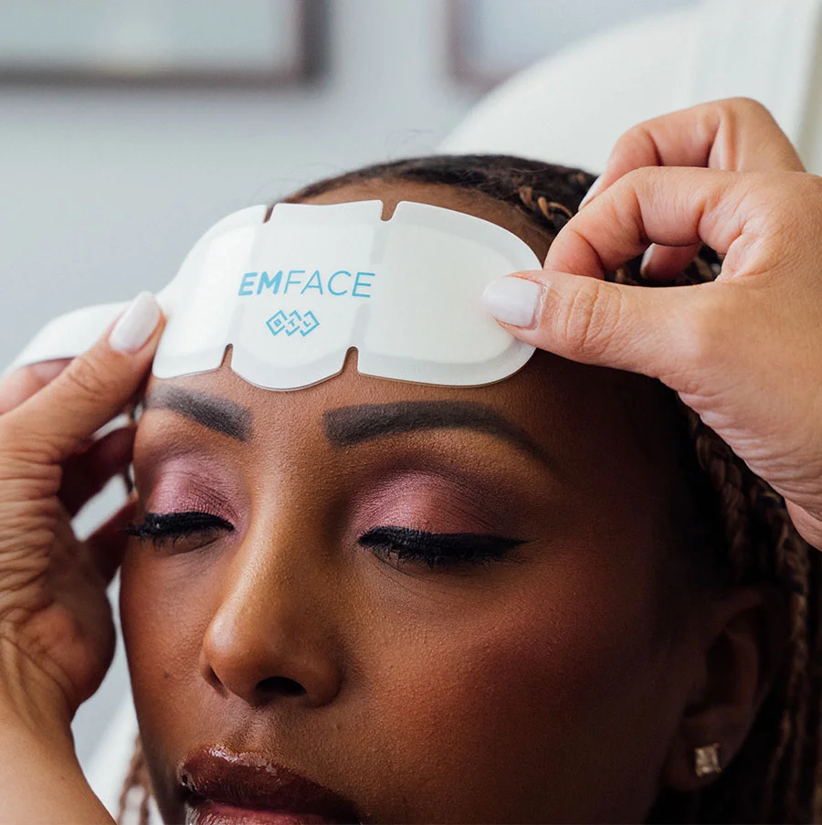 A woman with dark skin and braided brown hair wears bold makeup—purple eyeshadow, dark eyeliner, and red lipstick. She adjusts a white “EMFACE” cloth on her forehead with white-painted nails. A diamond-shaped earring and soft lighting frame her face, while a blurred medspa-like background suggests a beauty or facial treatment scene. - EM Face Benefits in San Jose, CA
