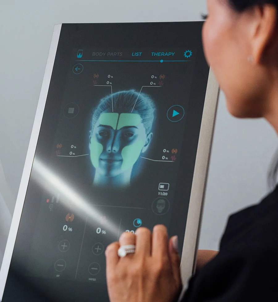 A close-up shot of a woman's hand touching the screen of a modern medical or aesthetic device. The screen displays a glowing blue digital representation of a female face with sections highlighted in green, along with technical menus, therapy options, and percentage indicators for a treatment like EMface. The woman is wearing a ring and a black shirt. - EM Face in San Jose, CA
