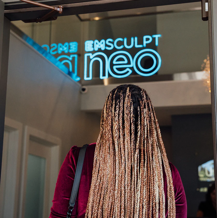 A rear view of a person with long, blonde and brown box braids, wearing a deep red velvet top, walking towards a glass door. The door has the words "EMSCULPT neo" prominently displayed in bright, neon blue light. The image suggests entry into a clinic or treatment center. - Emsculpt Neo Benefits in San Jose, CA