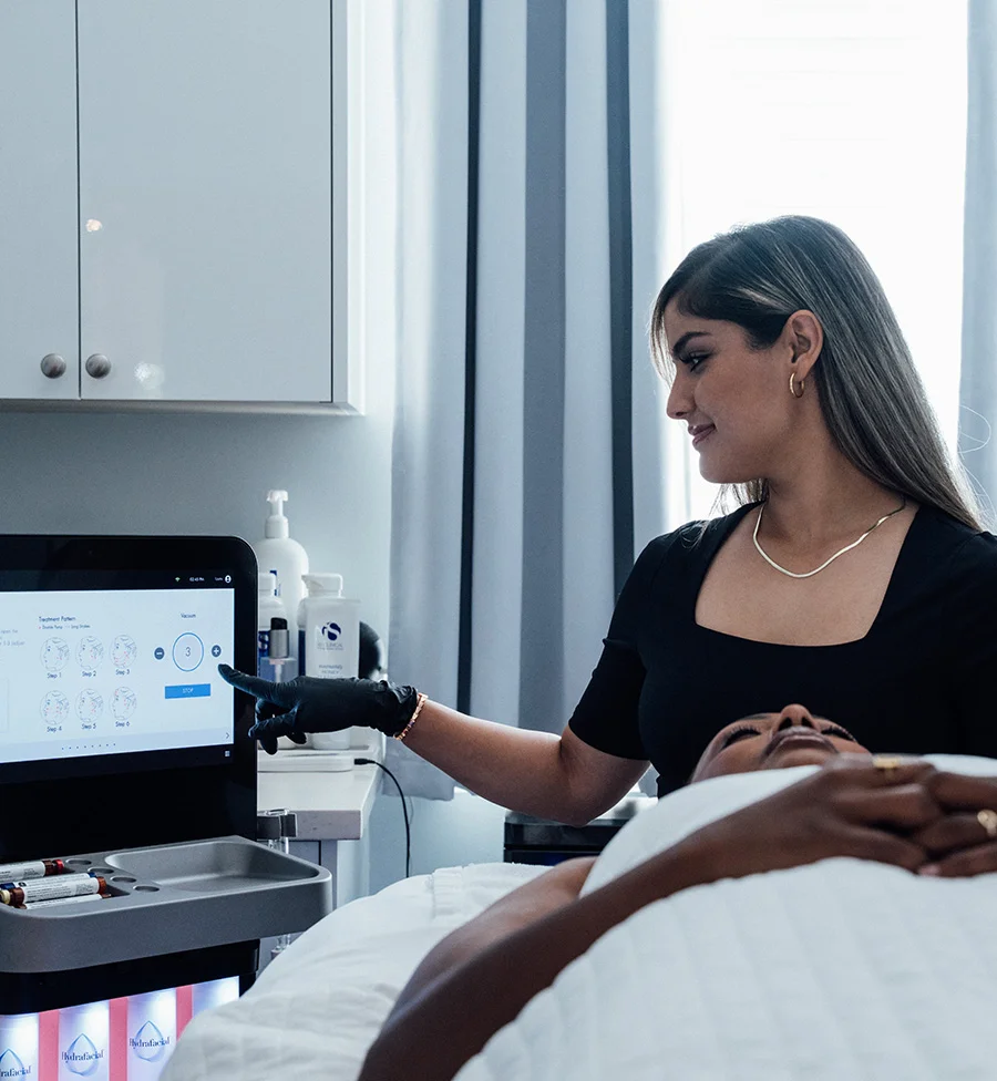 A spa or clinic scene where a professional with long hair and a black top is pointing to a screen displaying a treatment protocol, likely for a HydraFacial, on a machine. The client is lying on a treatment bed, covered with a white sheet, in the foreground. The professional is wearing black gloves and discussing the steps with the client. - Hydrafacial Canidates in San Jose, CA