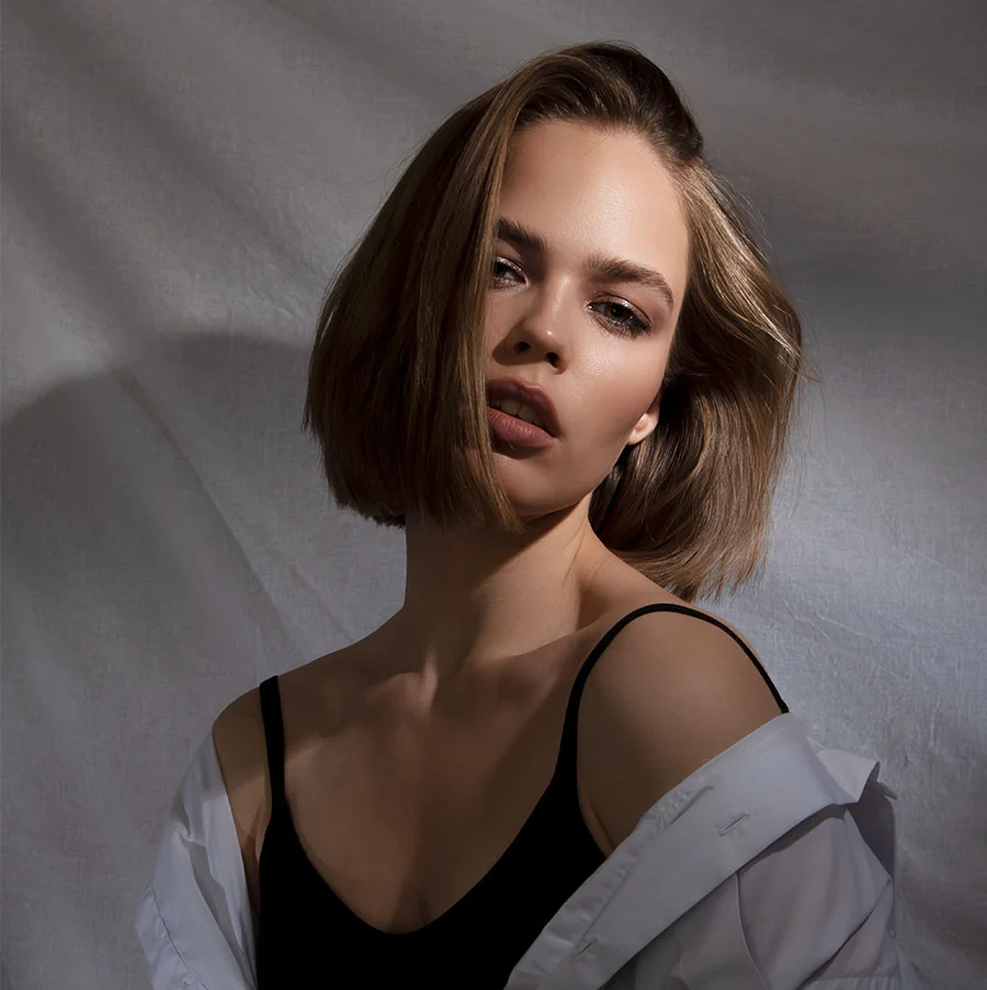 A portrait of a young woman with a short bob haircut and a confident expression looking directly at the camera. She is wearing a black tank top under an unbuttoned white collared shirt that is falling off one shoulder. The lighting is dramatic, casting shadows and highlights on her face, and the background is a white, crumpled or draped fabric. - IPL Benefits in San Jose, CA