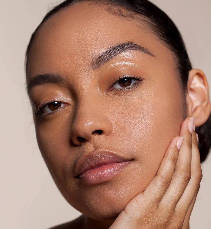 A close-up portrait of a young woman with smooth, radiant brown skin, looking directly at the camera. She has her hand gently resting on her chin and cheek. Her hair is pulled back, highlighting her high cheekbones and well-groomed eyebrows. The background is a soft, light beige. - IPL Canidates in San Jose, CA