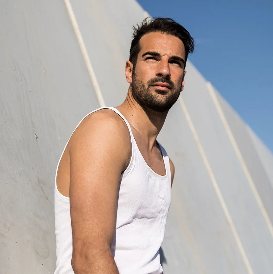 An outdoor portrait of a man with dark hair and a beard, wearing a white tank top. He is leaning against a textured light gray or white wall, looking up and to the side. The scene is brightly lit by the sun, casting a shadow and highlighting the clear blue sky in the background. - IV Therapy in San Jose, CA