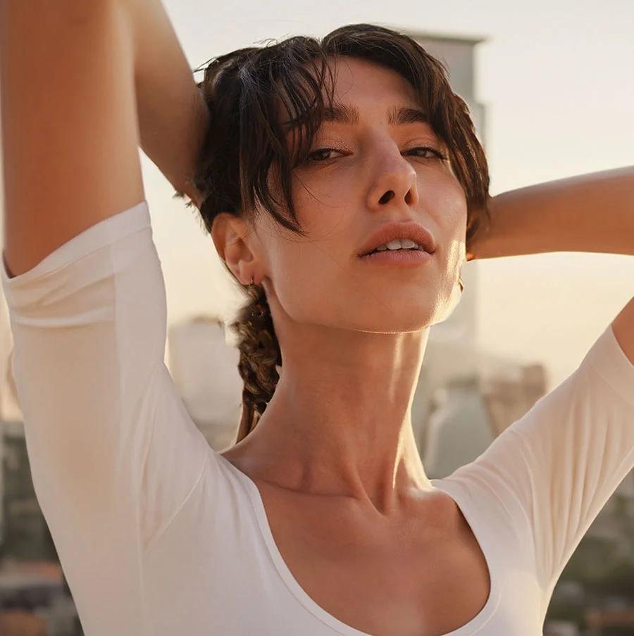 A woman with shoulder-length, dark brown hair is seen from the mid-chest up. She is wearing a off-white, loose-fitting blouse with short sleeves. Her hands are resting on her head, and her expression is neutral, though slightly directed toward the viewer. The lighting suggests a golden hour, with a soft, warm glow. Her skin appears smooth and light-toned.- Kybella in San Jose, CA