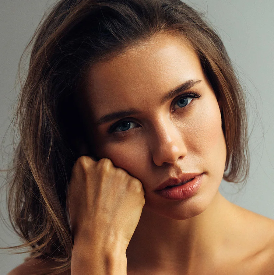 A close-up headshot of a young woman with a direct, serious expression looking at the camera. She has wavy brown hair, striking blue eyes, and is touching her chin with a clenched fist. Her bare shoulders are visible, and the background is a solid, light gray. - Laser Scar Removal in San Jose, CA