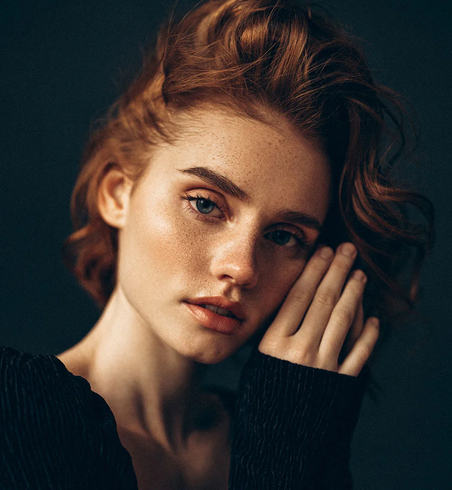 A close-up dramatic portrait of a young woman with fair skin, freckles, and reddish-auburn hair. She is looking directly at the camera with an intense expression and has one hand resting against her cheek. She is wearing a dark, ribbed top, and the background is a solid, dark gray or black. - Laser Scar Removal in San Jose, CA