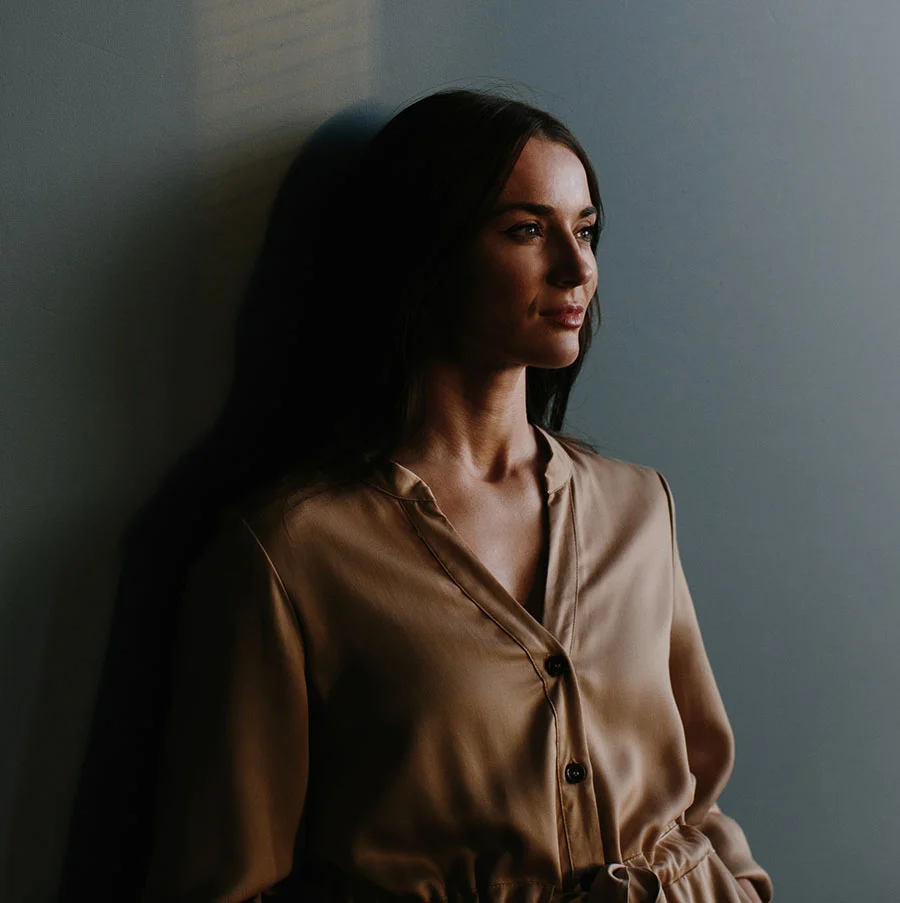 A portrait of a woman with long, dark hair, wearing a silky, light brown or tan collared shirt with buttons. She is leaning against a muted blue-gray wall in a dimly lit room, with a soft light illuminating her face from the side as she looks off-camera. - Laser Skin Resurfacing in San Jose, CA
