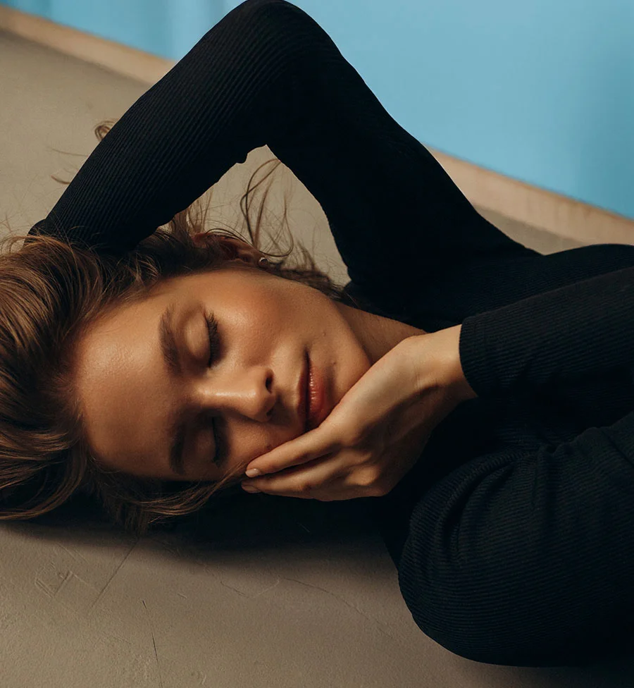 A close-up artistic shot of a woman with her eyes closed, lying on a light gray surface. She is wearing a black ribbed top, with her arms above her head and one hand touching her cheek. Her face is softly illuminated, and the background behind her head is a solid blue. - Laser Skin Resurfacing in San Jose, CA
