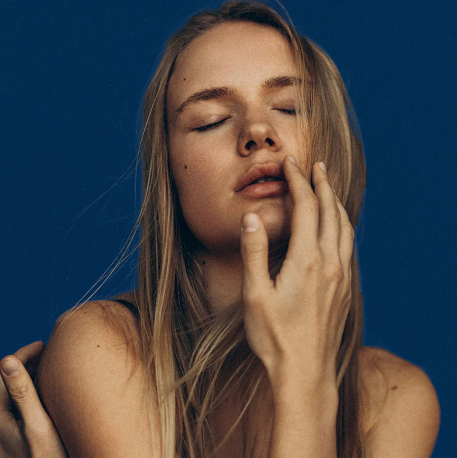 A close-up artistic portrait of a young woman with long blonde hair and fair skin. Her eyes are closed, and she has one hand gently touching her chin and lower lip. A second hand rests on her shoulder, suggesting an embrace or close interaction. The background is a solid, deep blue. - Lip Fillers in San Jose, CA