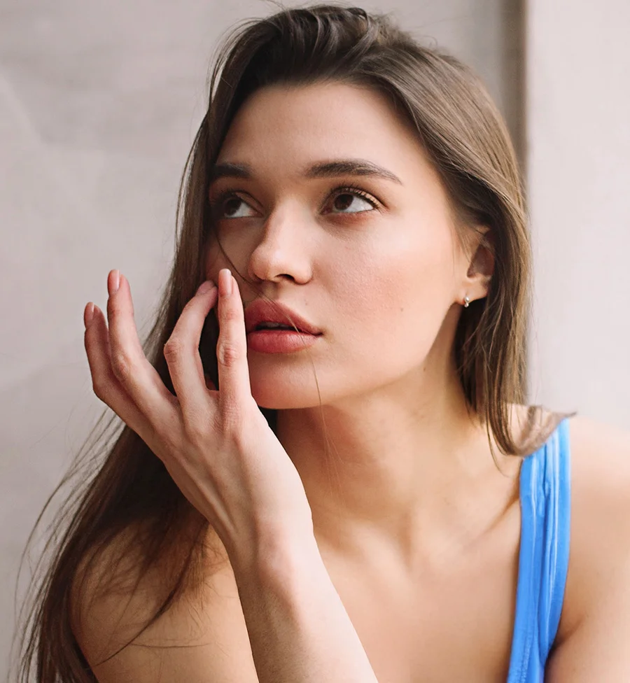 A portrait of a young woman with long brown hair, wearing a blue strap top, looking up and to the side. She is touching her face near her mouth and chin with her right hand, with a thoughtful or pensive expression. The background is a plain light gray wall. - Lip Fillers in San Jose, CA