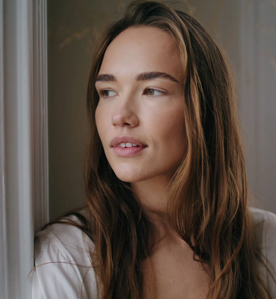 A close-up portrait of a young woman with long, brown, wavy hair looking off to the side, leaning against a white window frame in a softly lit room. She has clear skin, full lips, and natural-looking makeup. The lighting suggests she is near a window with a muted, warm-toned background. - Microneedling in San Jose, CA