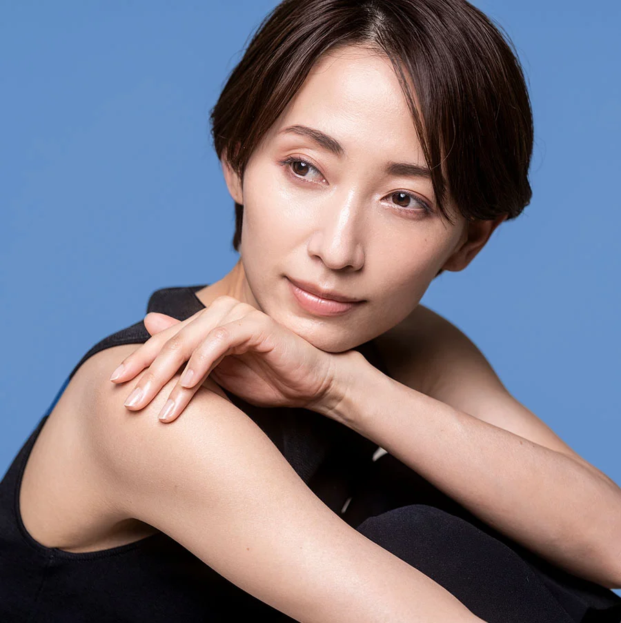 The image is a professional portrait of a woman with short dark hair styled sleekly, wearing a black tank top. She sits cross-legged, her right arm resting on her knee with her hand under her chin, creating a poised and elegant pose. Set against a solid blue background, the portrait conveys sophistication, confidence, and professionalism - O Shot in San Jose, CA