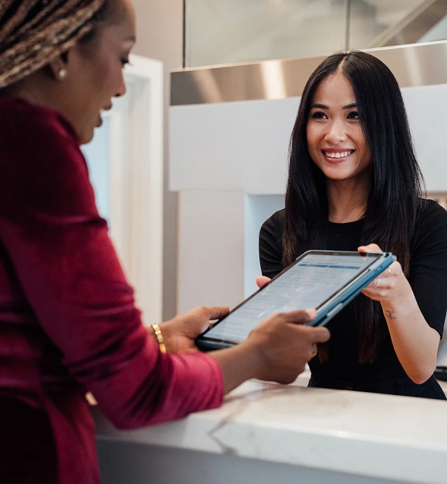 A woman with yellow braids wearing a red blouse and gold jewelry hands a tablet to another woman with long black hair seated behind a white counter. The second woman, dressed in black, takes the device as a large opening framed in silver metal and a brushed silver countertop define the background. The beige walls and service-style setup suggest an office or customer-service location. - O Shot in San Jose, CA