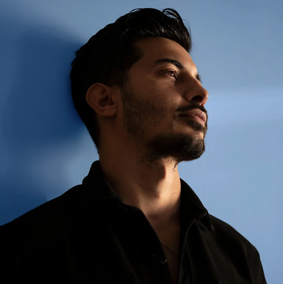 A man with dark, spiky hair and a full, trimmed beard gazes upward with a calm, introspective expression. He wears an open-collared black button-up shirt, its shadows adding depth and contrast. Positioned on the right side of the frame against a light blue-gray wall, his tilted head and slightly furrowed brow suggest a candid moment of quiet contemplation.- P Shot in San Jose, CA