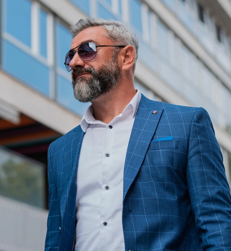 A gray-haired man with a neatly trimmed beard stands confidently in front of a multi-story building under a bright daytime sky. He wears a blue suit jacket with white grid lines, a white collared shirt, and a blue pocket square, complemented by stylish sunglasses. The blurred background suggests an office or residential building, while his polished appearance conveys professionalism and sophistication. - P Shot in San Jose, CA