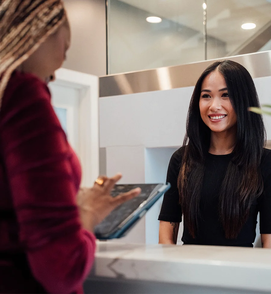A photo taken in a modern clinic or reception area. A friendly woman with long dark hair, wearing a black shirt, is standing behind a white counter, smiling and facing a client. The client, who has blonde braids and is wearing a maroon outfit, is partially visible from the back and is holding a tablet. - PDO Threads in San Jose, CA