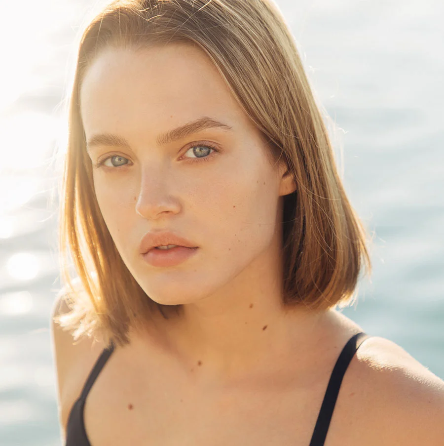 A close-up outdoor portrait of a young woman with a short, light brown bob haircut and bright blue eyes, looking directly at the camera. She is wearing a black tank top. Bright sunlight from behind and to the side creates a glowing halo effect around her hair, and the background is a blurred body of water with sunlight reflecting off the surface. - Picosure Pro in San Jose, CA