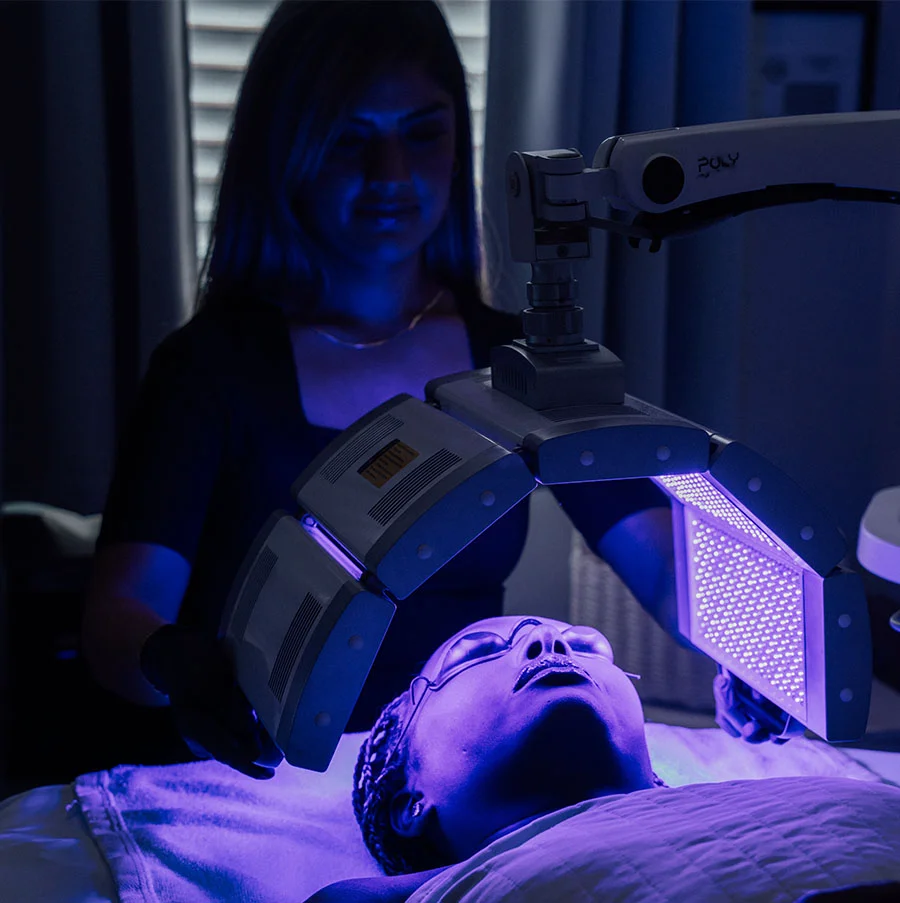 A woman is receiving an LED light therapy treatment from an arched, mechanical device, labeled "POLY," which is emitting a bright **purple or violet light** over her face and chest. She is lying on a bed with protective eyewear on. A technician, wearing a black top and black gloves, stands behind the device, observing the procedure in the dimly lit treatment room. - Poly Led Light Therapy in San Jose, CA