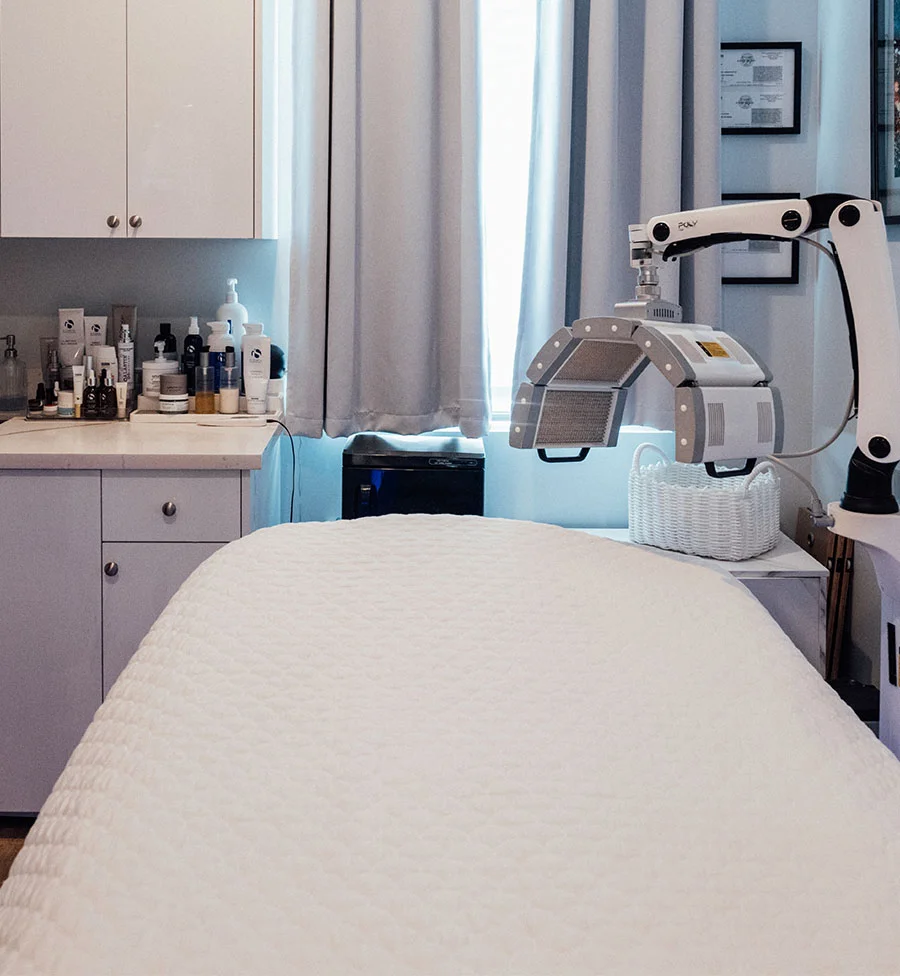 A photo of a brightly lit, clean treatment room, likely in a spa or clinic. In the center is a white treatment bed covered with a quilted sheet. Above the bed is a freestanding, arched LED light therapy machine, labeled "POLY". To the left is a white cabinet and counter with various skincare products, and the background features light-colored curtains over a window. - Poly Led Light Therapy in San Jose, CA