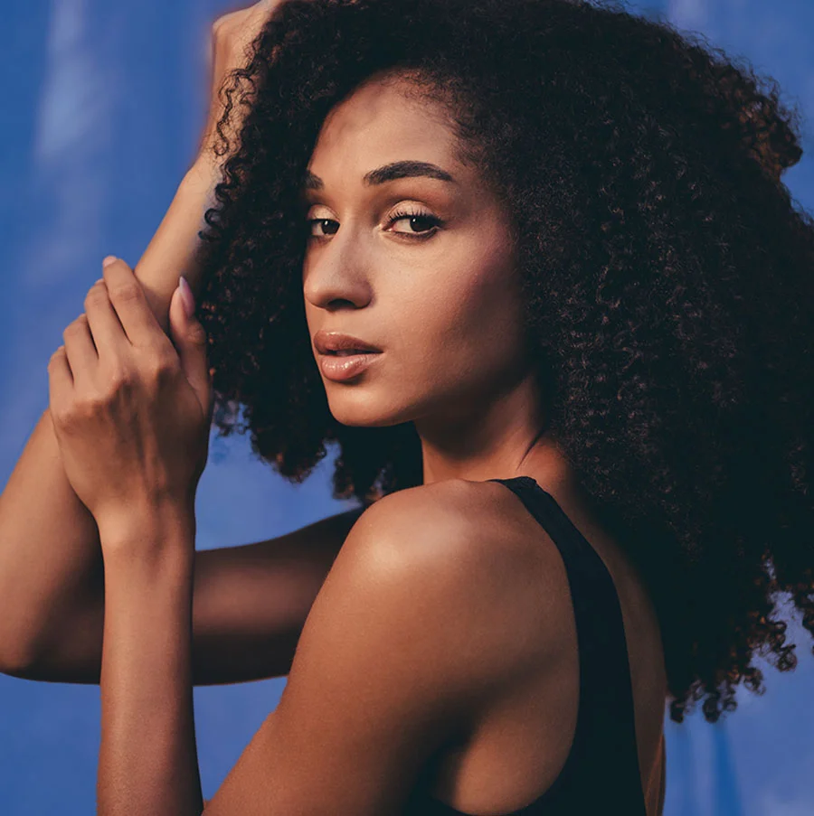A close-up artistic portrait of a woman with dark, voluminous curly hair, looking over her shoulder directly at the camera. She is wearing a black tank top, and her arms are raised, framing her head. The lighting highlights her features, and the background is a solid, vibrant blue. - PRP for Sexual Health in San Jose, CA