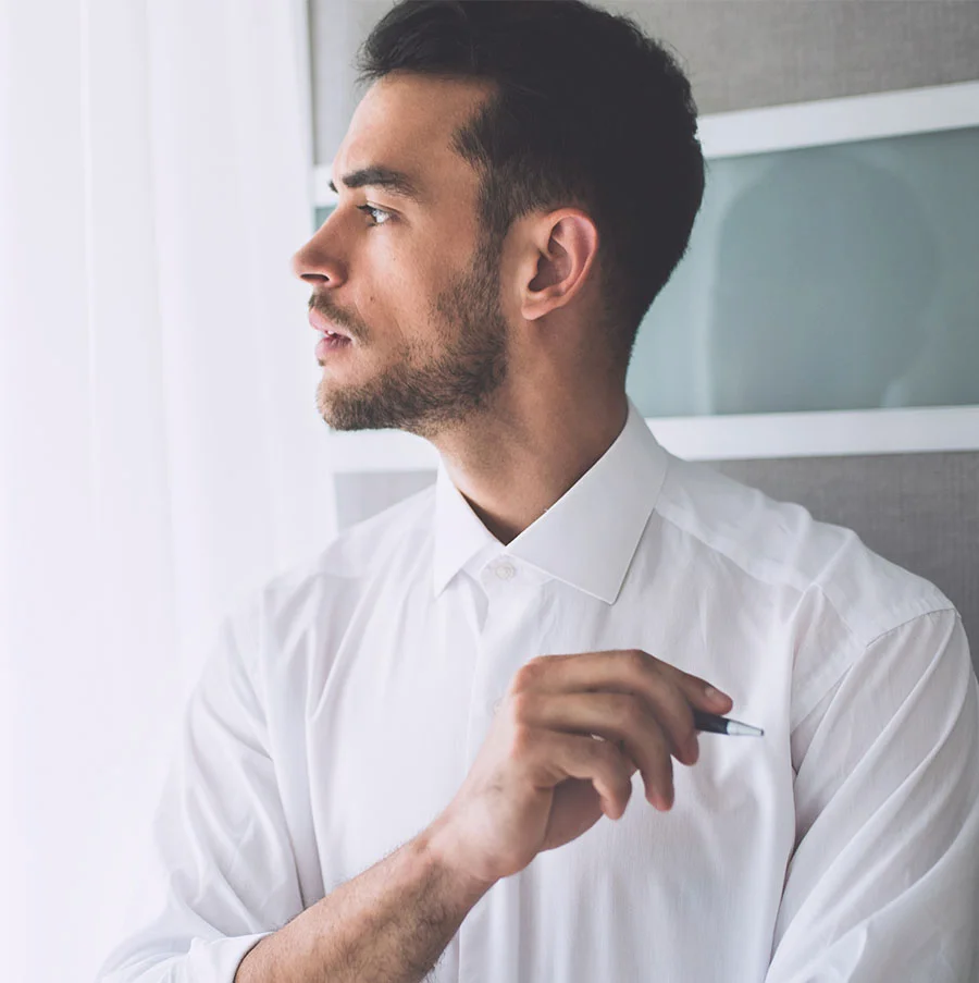 A portrait of a man with dark hair and a beard, wearing a white collared shirt, looking off to the side. He is standing near a window with bright light coming in, and he is holding a small dark pen or object in his right hand. The background includes a muted gray wall or door. - PRP Hair Restoration in San Jose, CA