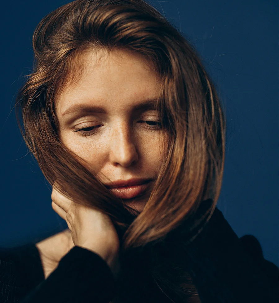 A close-up artistic portrait of a woman with reddish-brown hair and freckles, looking down with her eyes closed or partially closed. Her hair is slightly windswept, covering part of her face, and one hand is resting gently against her cheek. She is wearing a dark top, and the background is a solid, dark blue. - RPP Hair Restoration in San Jose, CA