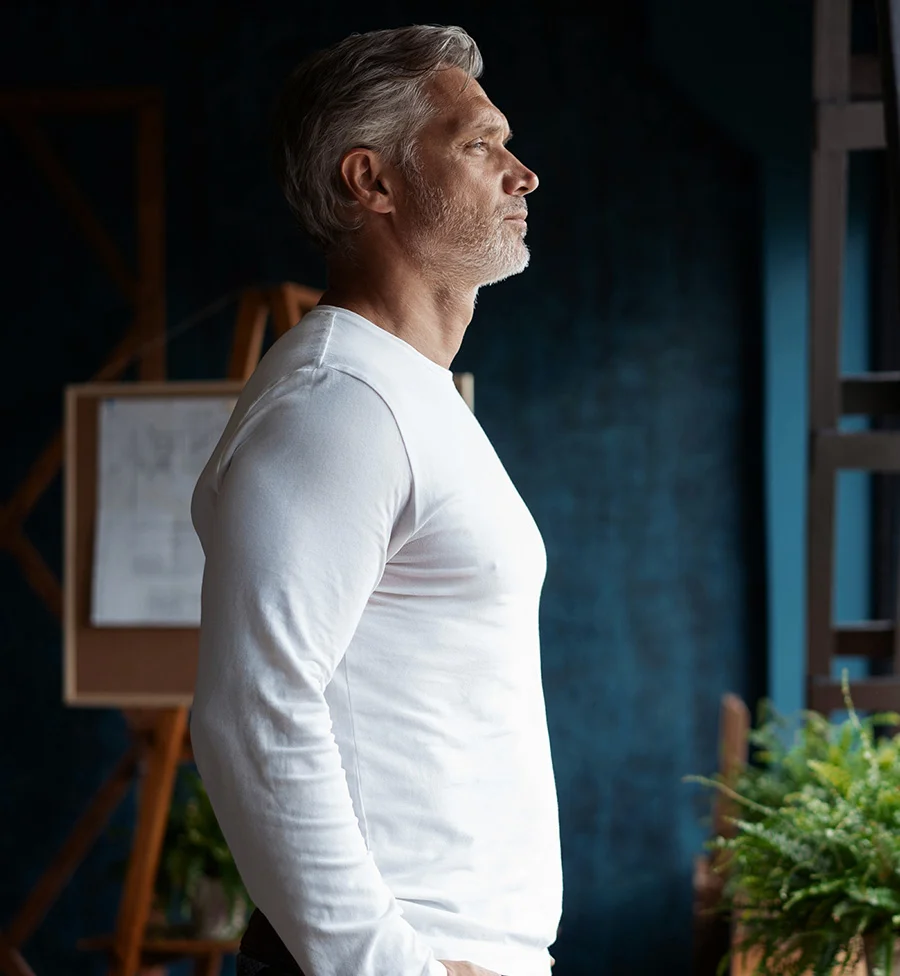 A side profile portrait of a mature man with a distinguished gray beard and white hair, looking contemplatively out a window. He is wearing a long-sleeved white shirt and is standing in a room with a dark blue wall. A white easel and a green potted plant are visible in the background. - PRP Stem Cells for Joint Repair in San Jose, CA