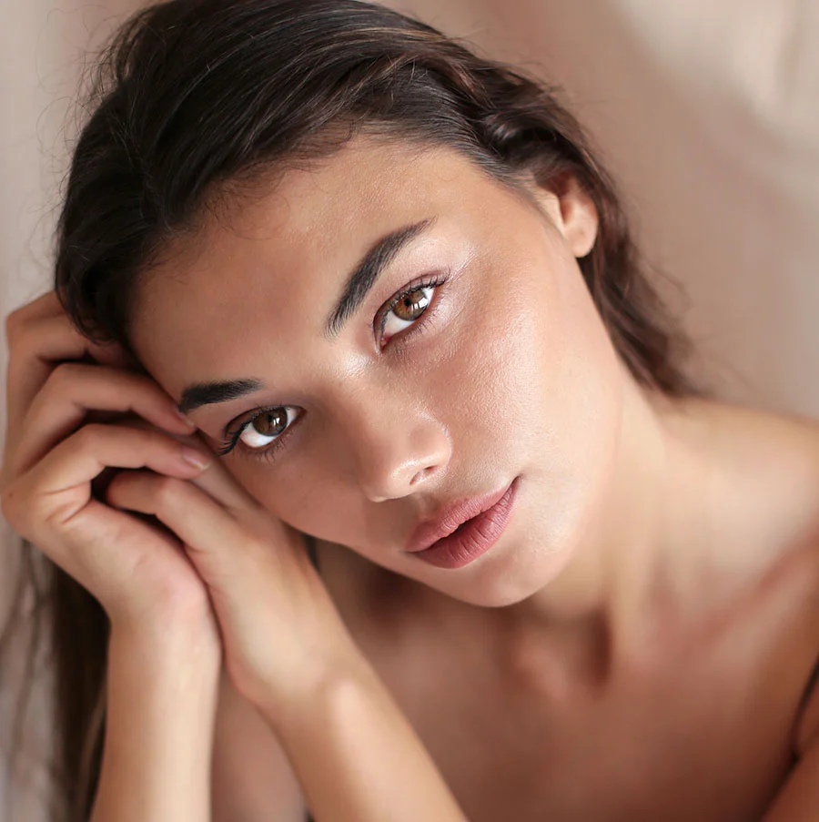 A close-up, high-angle portrait of a woman with long, dark hair and striking brown eyes looking directly at the camera. Her hands are gently clasped near her face, and her shoulders are bare, suggesting an intimate or artistic pose. The lighting is soft and even, and the background is a muted, light beige. - Rejuran in San Jose, CA