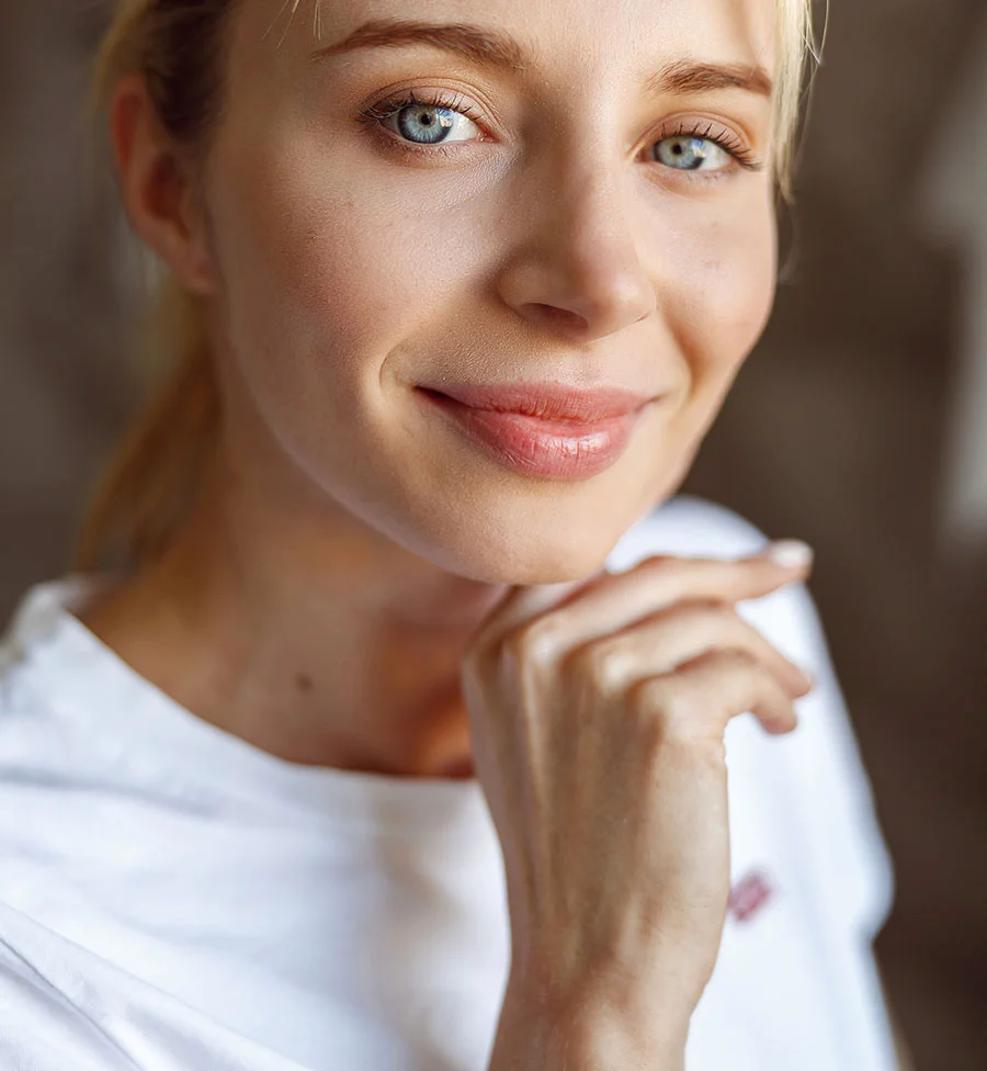 A close-up portrait of a young woman with light blonde hair, bright blue eyes, and a pleasant smile. She is looking directly at the camera, wearing a white t-shirt, and has one hand gently touching her chin. The background is a blurred, warm brown color. - Rejuran in San Jose, CA