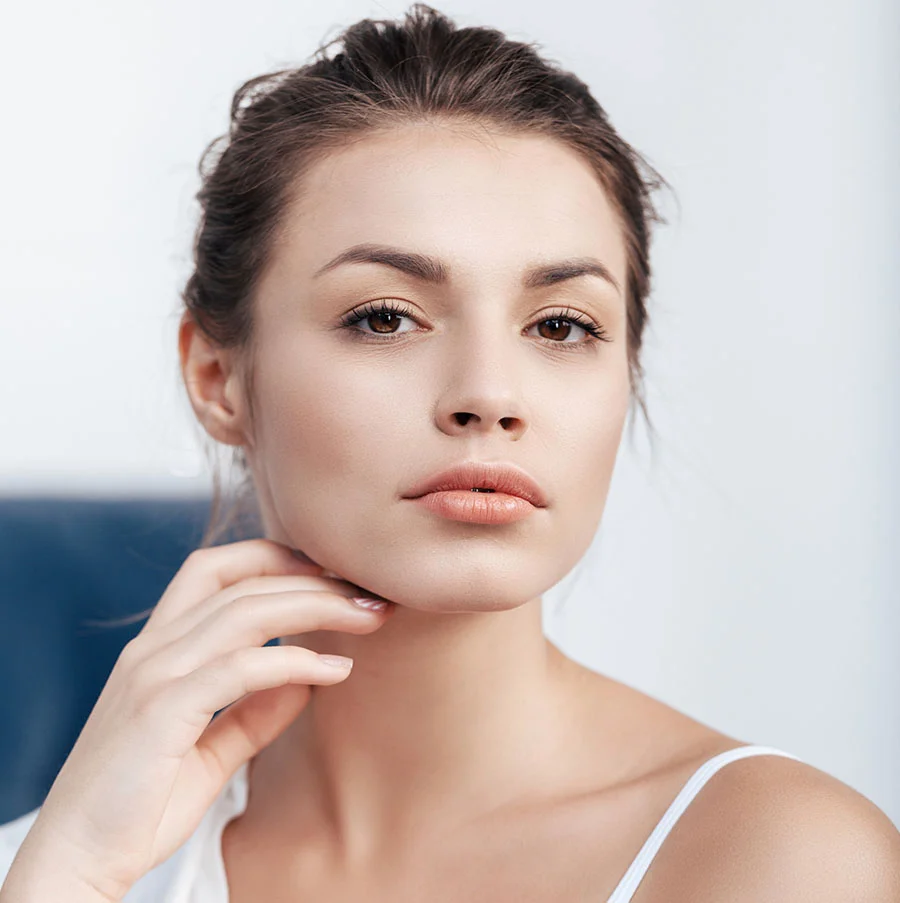 A close-up portrait of a young woman with her hair pulled back, looking directly at the camera with a neutral expression. She is wearing a white tank top and gently touching the underside of her jaw with one hand. The background is softly lit, with a hint of a dark blue shape to the left. - RF Microneedling in San Jose, CA