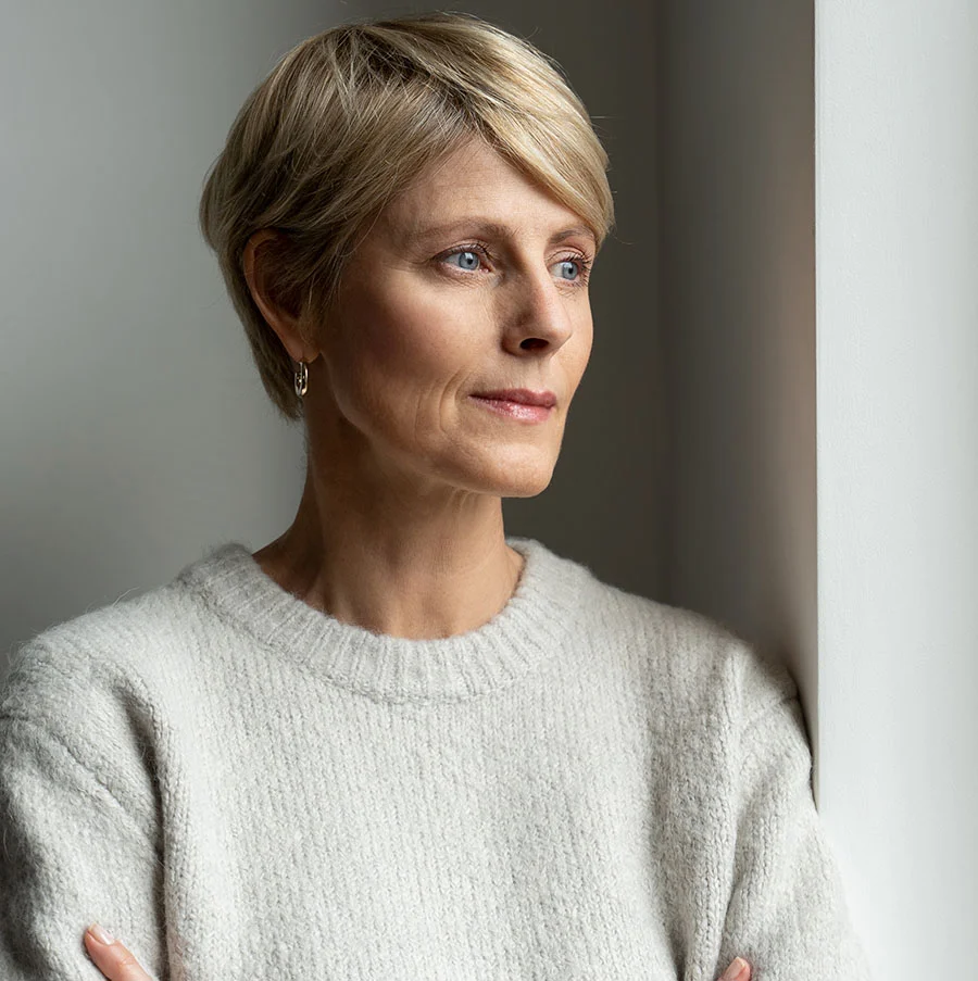 A professional portrait of a woman with short blonde hair in a neat undercut, light brown brows, and blue eyes looking right. She wears silver-tone hoop earrings and a soft white knit sweater. Her fair skin has a light pink hue. She leans an arm along a textured white wall, which creates a soft, balanced backdrop. Her expression is neutral and thoughtful. - Stem Cell Concierge in San Jose, CA