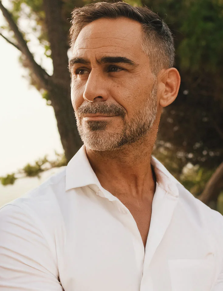 Close-up of a man in a white button-down shirt, facing left with a faint smile. He has tan skin, gray-streaked stubble, and brown eyes. Blurred greenery in the background suggests an outdoor setting. - Stem Cell Concierge in San Jose, CA