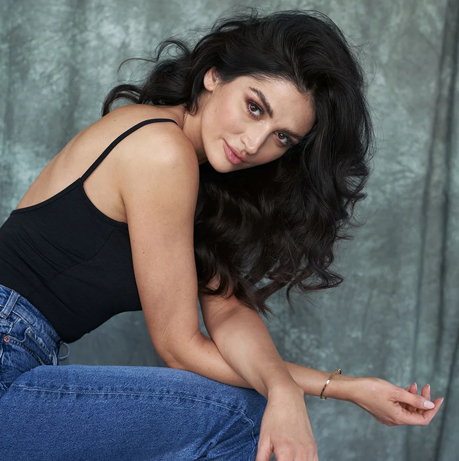 A woman with long, dark curly hair sits against a muted gray-blue textured wall. She wears a black sleeveless top and blue jeans, looking slightly to the left with a neutral, subtly suggestive expression. Soft lighting highlights her features, with the wall serving as a neutral backdrop. - Vaginal Rejuvenation in San Jose, CA