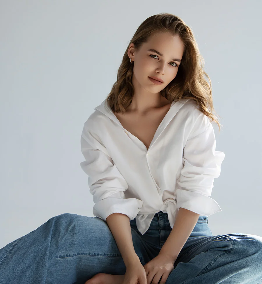 A young woman with wavy, light brown hair is sitting cross-legged, looking directly at the camera with a neutral expression. She is wearing a loose-fitting white collared shirt, unbuttoned at the top, and wide-leg blue jeans. The background is a plain, light gray or white. - Vaginal Rejuvenation in San Jose, CA