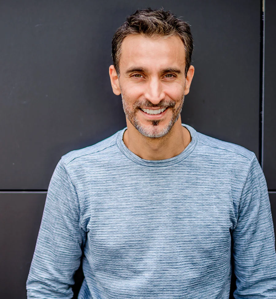 A portrait of a smiling middle-aged man with dark hair, a salt-and-pepper beard, and bright eyes. He is wearing a light blue, subtly textured long-sleeve shirt and is standing in front of a modern, dark gray or black paneled wall. - Vitamin Shot in San Jose, CA